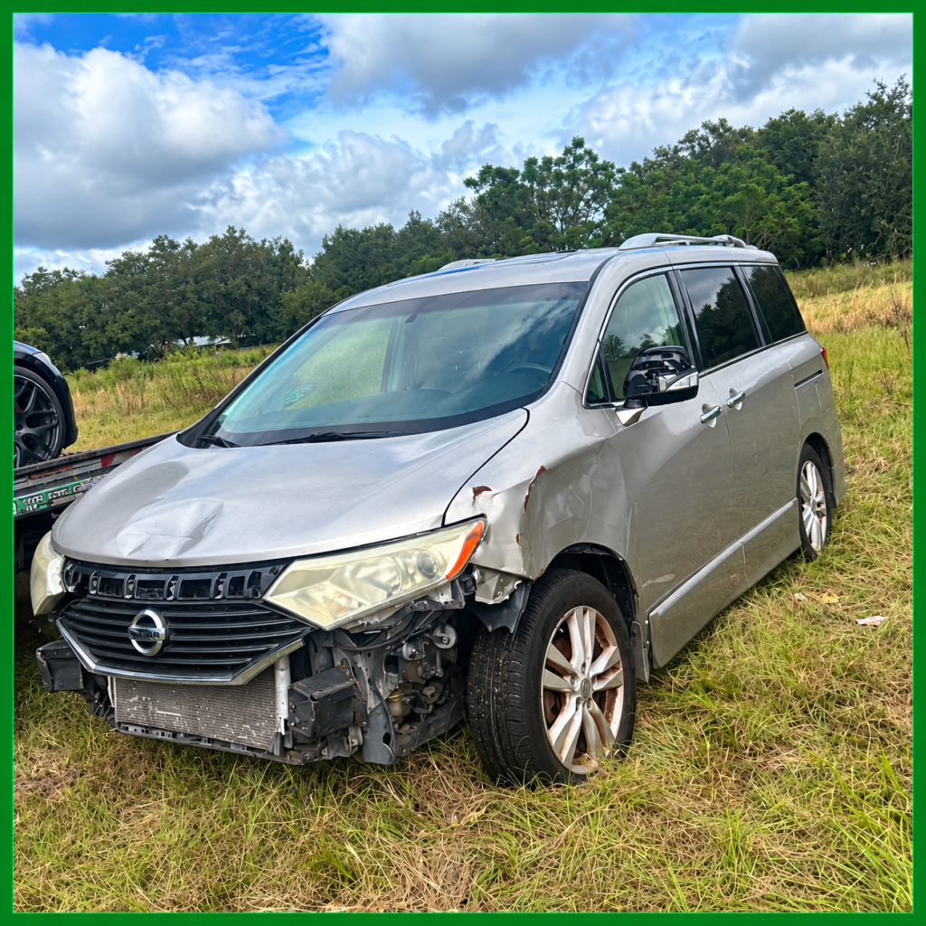 Damaged silver Nissan minivan with a missing front bumper in a grassy field